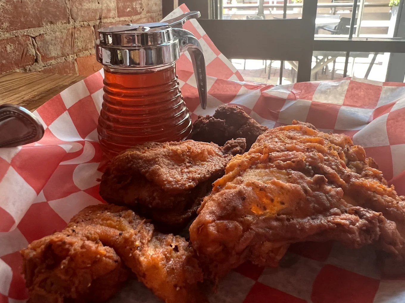 A basket of crispy fried chicken pieces sits on red and white checkered paper, next to a syrup dispenser, with sunlight streaming through a window in the background.