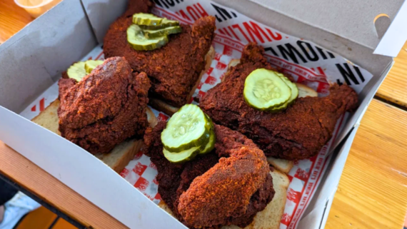 Four pieces of spicy fried chicken, each topped with pickle slices, are served on slices of white bread in a takeout box. The box sits on a wooden table, showcasing the mouthwatering flavors you&rsquo;d expect from top fried chicken restaurants.