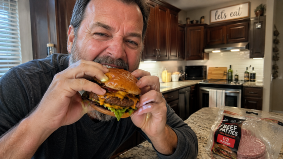 A man in a kitchen smiles and prepares to take a bite of a large cheeseburger. Packaged beef patties from popular frozen burger brands are visible on the counter next to him.