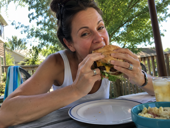 A woman wearing sunglasses on her head is about to take a big bite of a hamburger&mdash;possibly from one of her favorite frozen burger brands&mdash;while sitting at an outdoor table with food and drink on a sunny day.