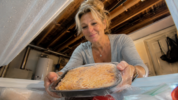 A woman smiles as she places one of her favorite frozen food products, a meal covered with cheese, into a chest freezer in a basement with exposed beams and a water heater in the background.