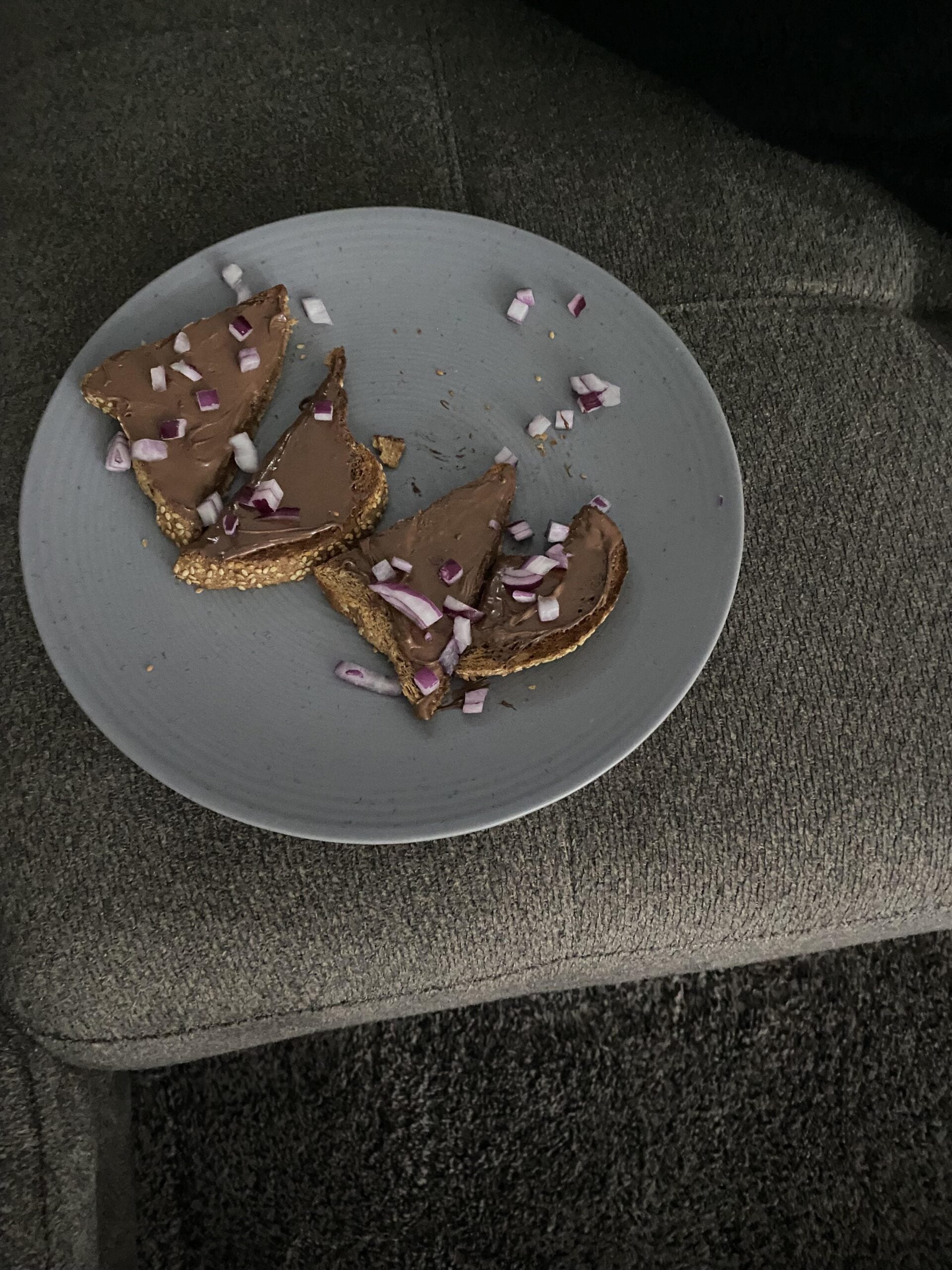 A grey plate with three slices of bread topped with a brown spread and chopped red onions, placed on a grey couch.