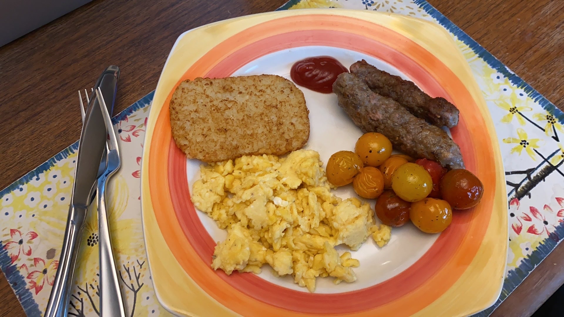 A plate with scrambled eggs, a hash brown, grilled sausages with ketchup, and yellow and red cherry tomatoes. Cutlery is placed on the left, and the plate sits on a colorful placemat with a floral design.