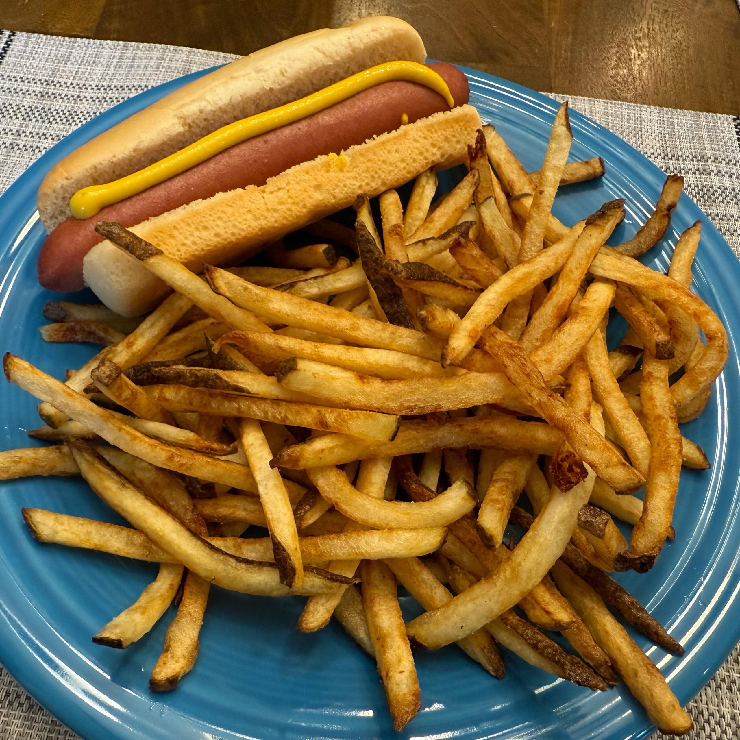 A blue plate holding a hot dog in a bun topped with mustard, served with a large pile of golden-brown French fries, sits on a woven placemat on a wooden table.
