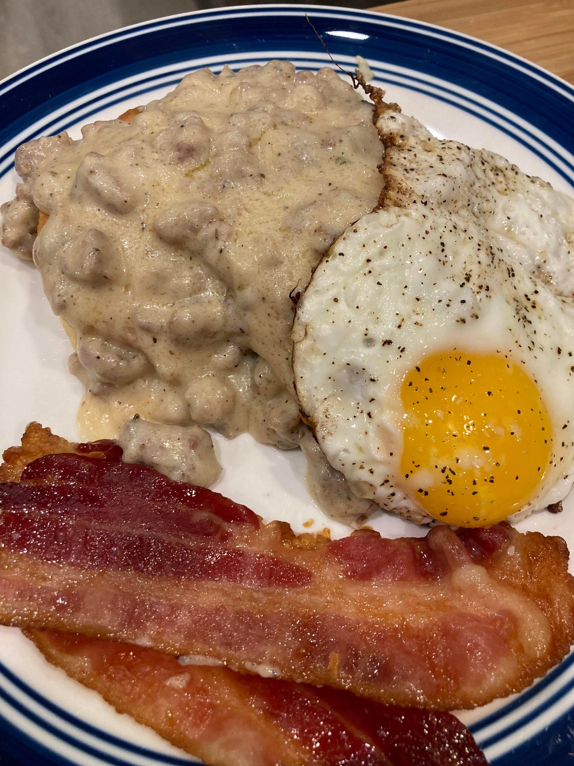 A plate with two strips of crispy bacon, a sunny-side-up egg, and a biscuit topped with creamy sausage gravy.