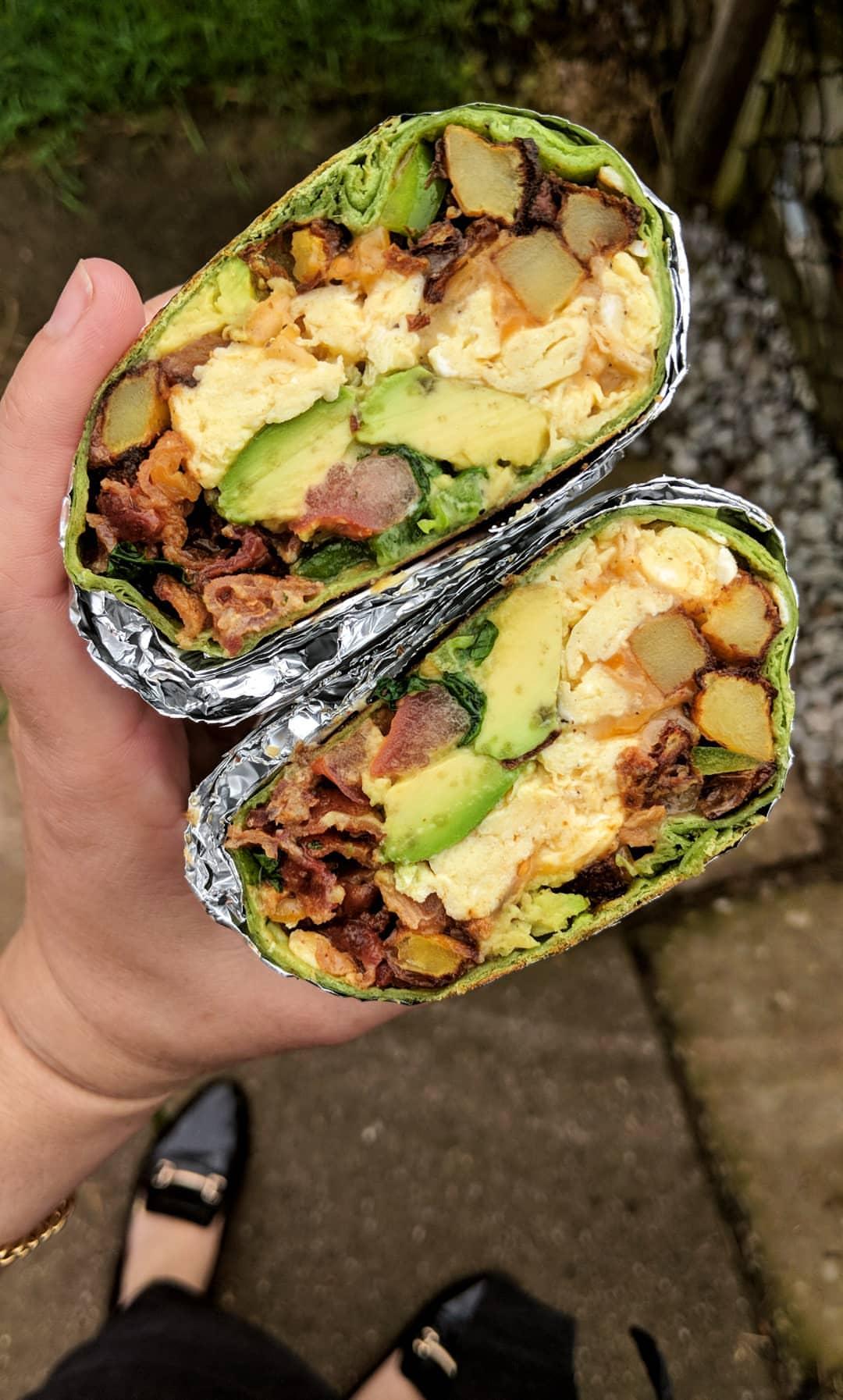 A hand holds a foil-wrapped breakfast burrito cut in half, showing eggs, avocado, potatoes, bacon, and spinach inside. Person stands on a sidewalk, wearing black shoes.