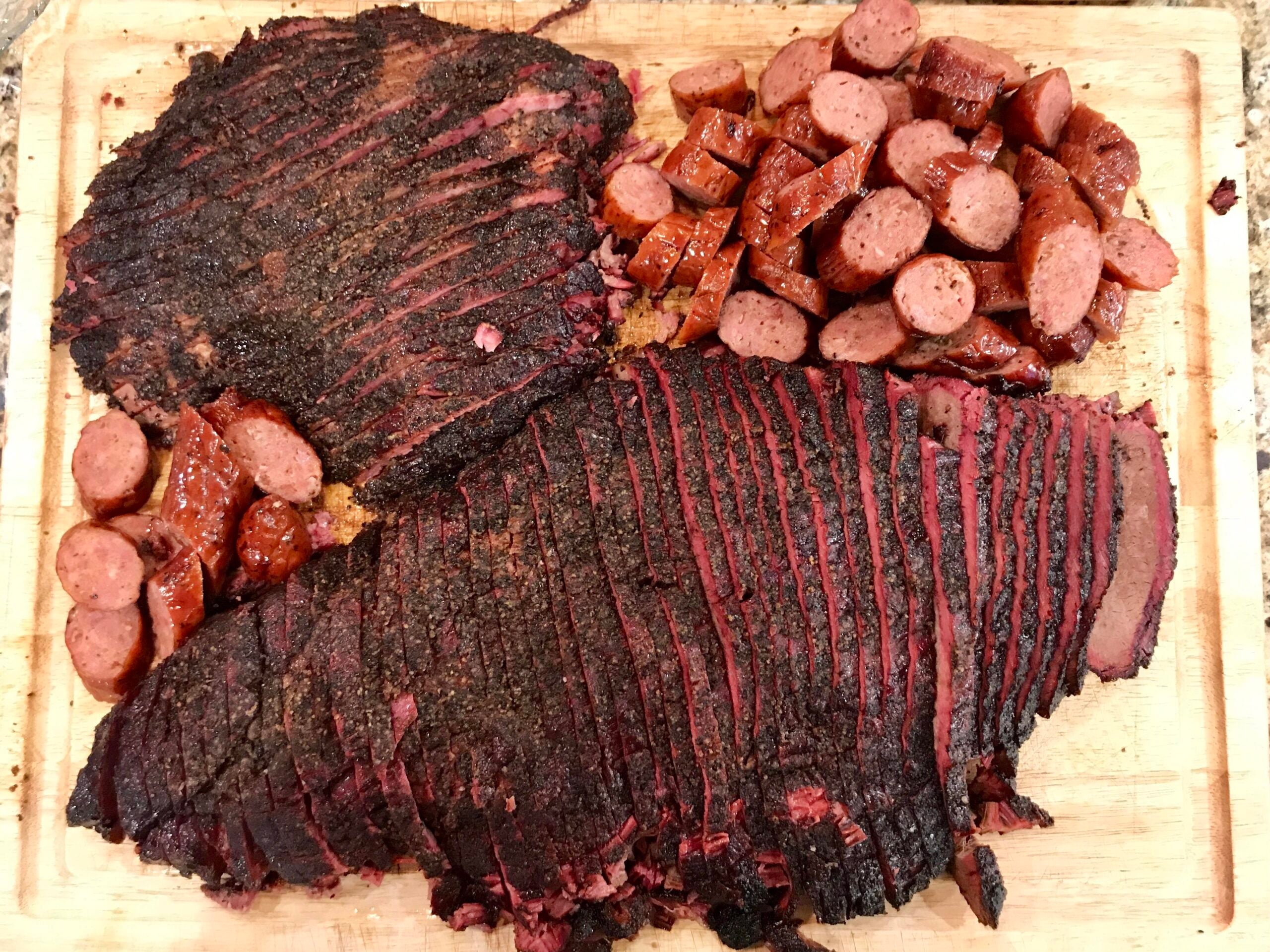 Sliced smoked brisket and pieces of sausage are arranged on a wooden cutting board, showing a dark, seasoned crust and juicy, tender interior.