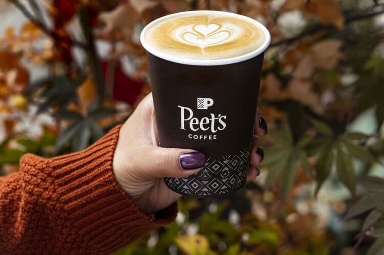 A hand with dark purple nail polish holds a Peet’s Coffee cup filled with latte art, capturing the cozy charm of coffee houses, while autumn leaves are blurred in the background.
