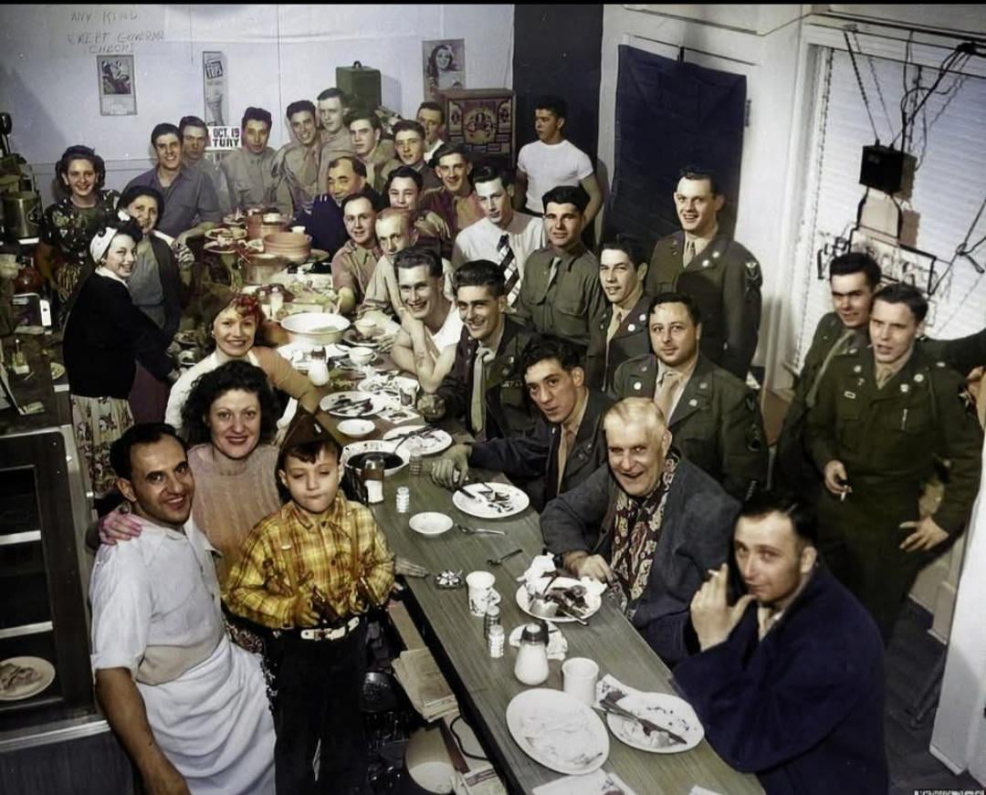 A large group of people, including men in military uniforms, women, and a child, gather around a diner counter for one of those nostalgic dinners, sharing food and smiling for a group photo.