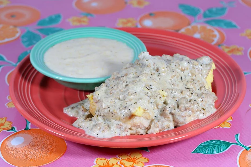 Best grits dishes. A plate with biscuits and eggs covered in creamy white sausage gravy, next to a bowl of grits, on a pink tablecloth with colorful fruit and floral patterns.