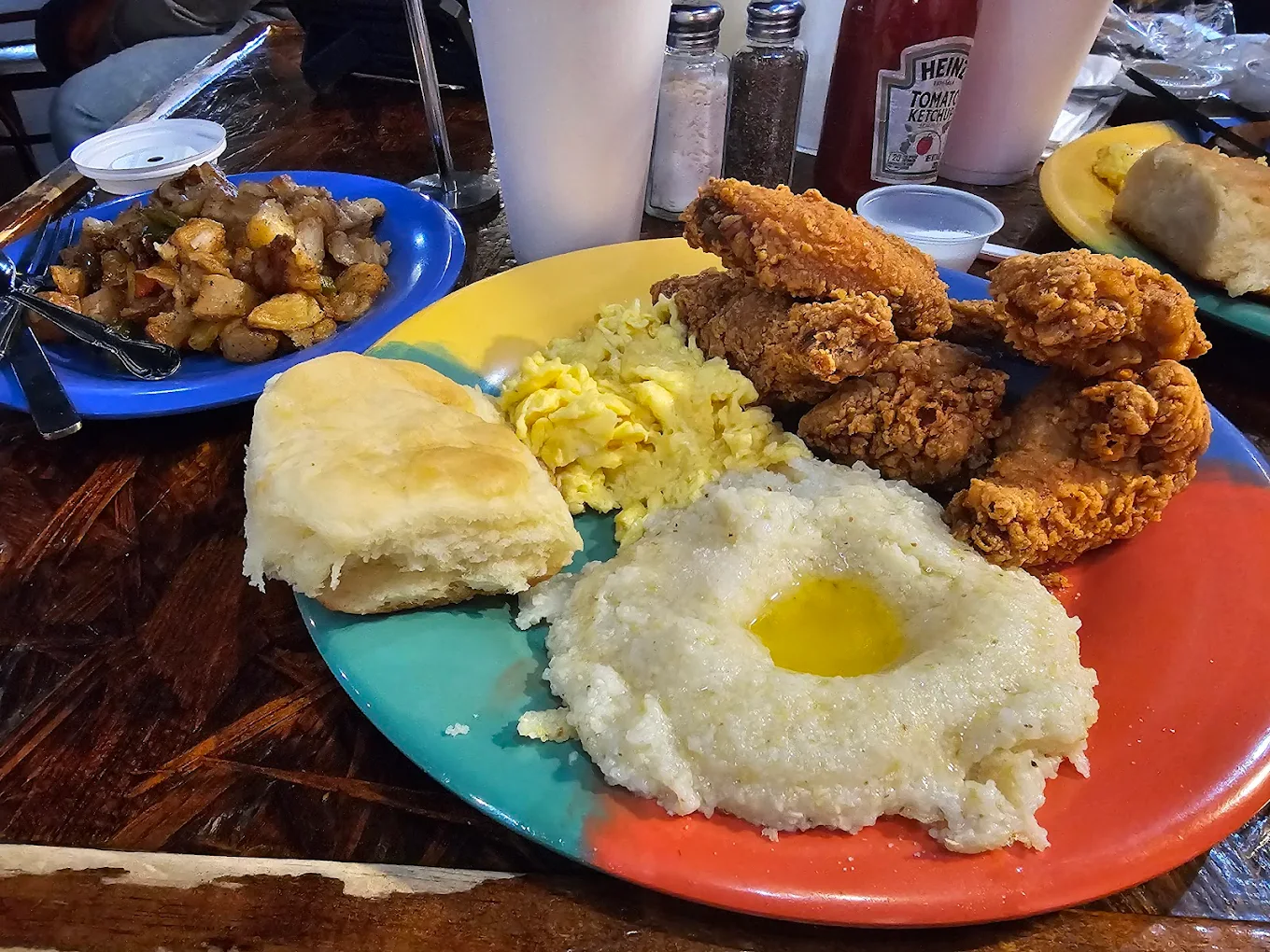 A colorful plate with fried chicken, scrambled eggs, creamy grits with melted butter, and a biscuit. On the side, a blue plate holds home fries. Drinks, condiments, and a biscuit are visible in the background.