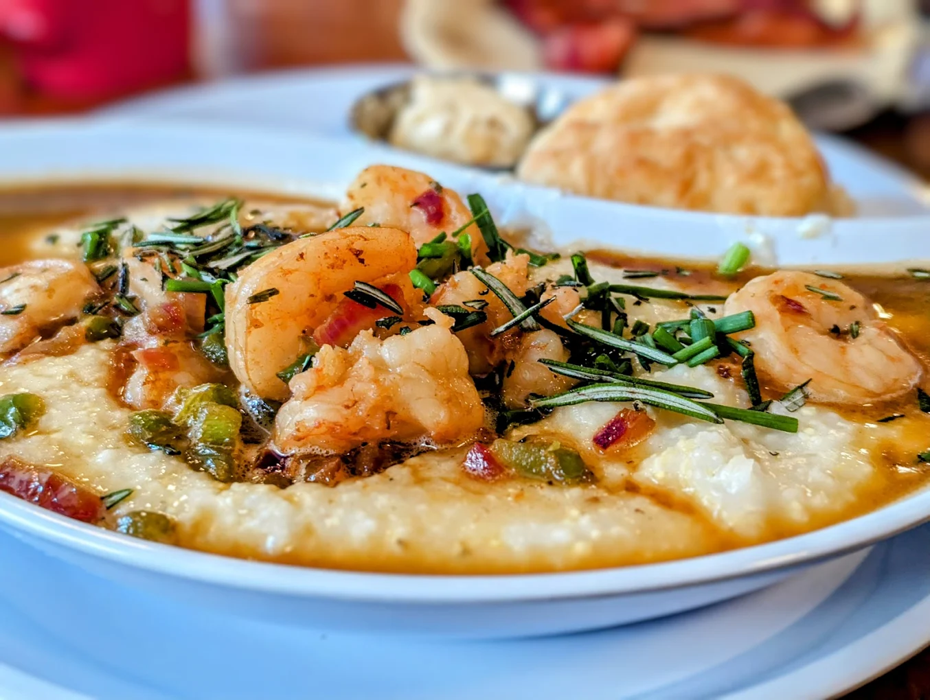 A close-up of shrimp and grits topped with chopped herbs on a white plate, with a biscuit and more food blurred in the background.