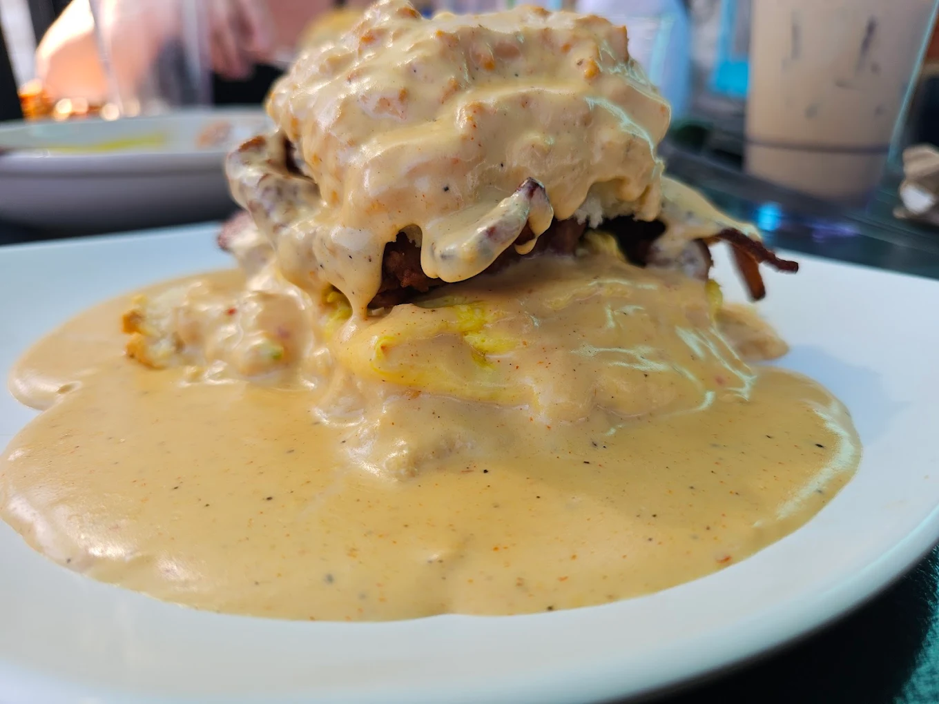 A close-up of a savory dish covered in creamy, pepper-speckled sauce on a white plate, with food layers barely visible under the thick sauce. Blurred background shows another plate and a beverage.