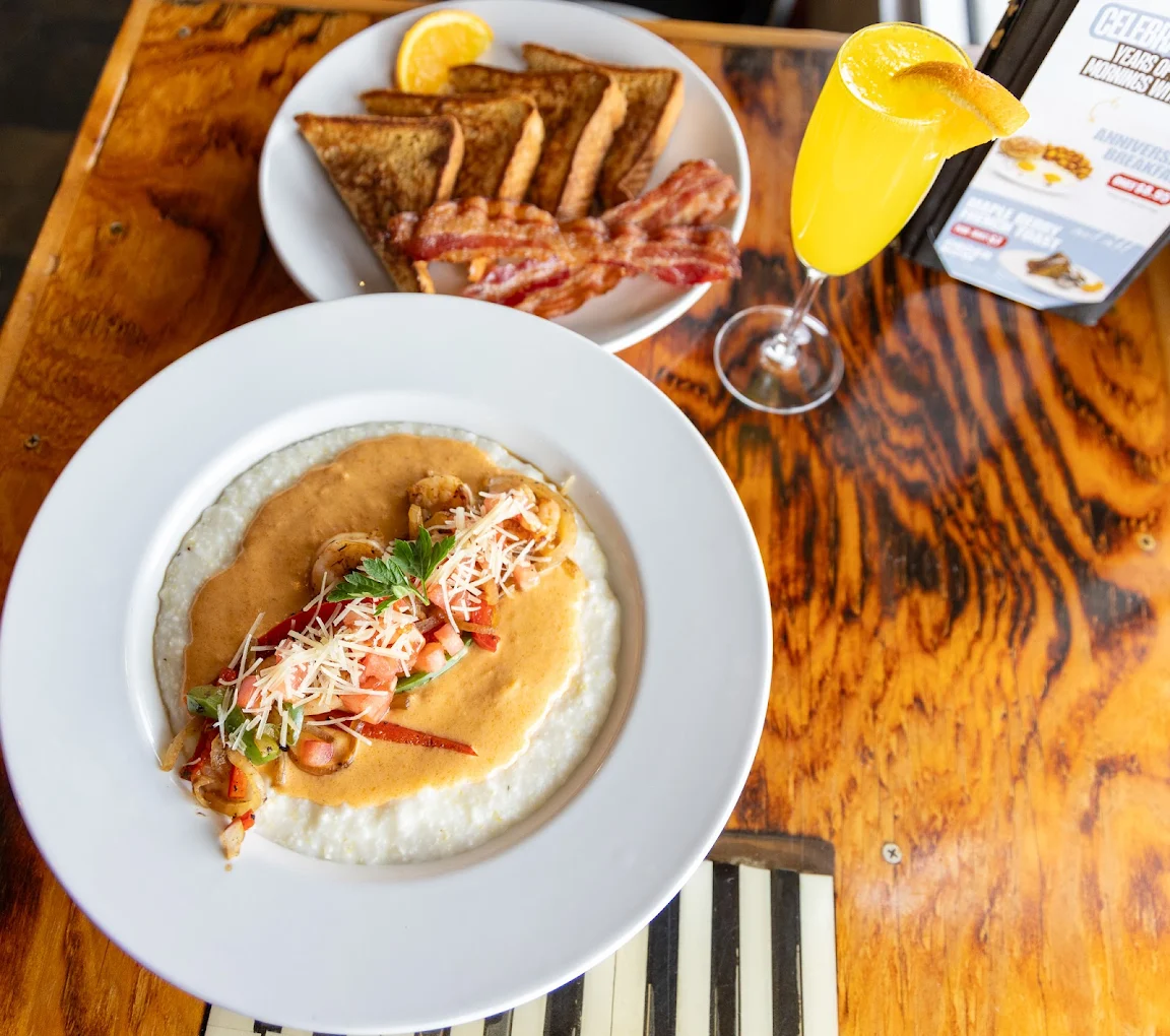 A plate of shrimp and grits topped with sauce and herbs sits next to a plate of toast, bacon, and orange slices, with a mimosa garnished with an orange wedge on a wooden table.