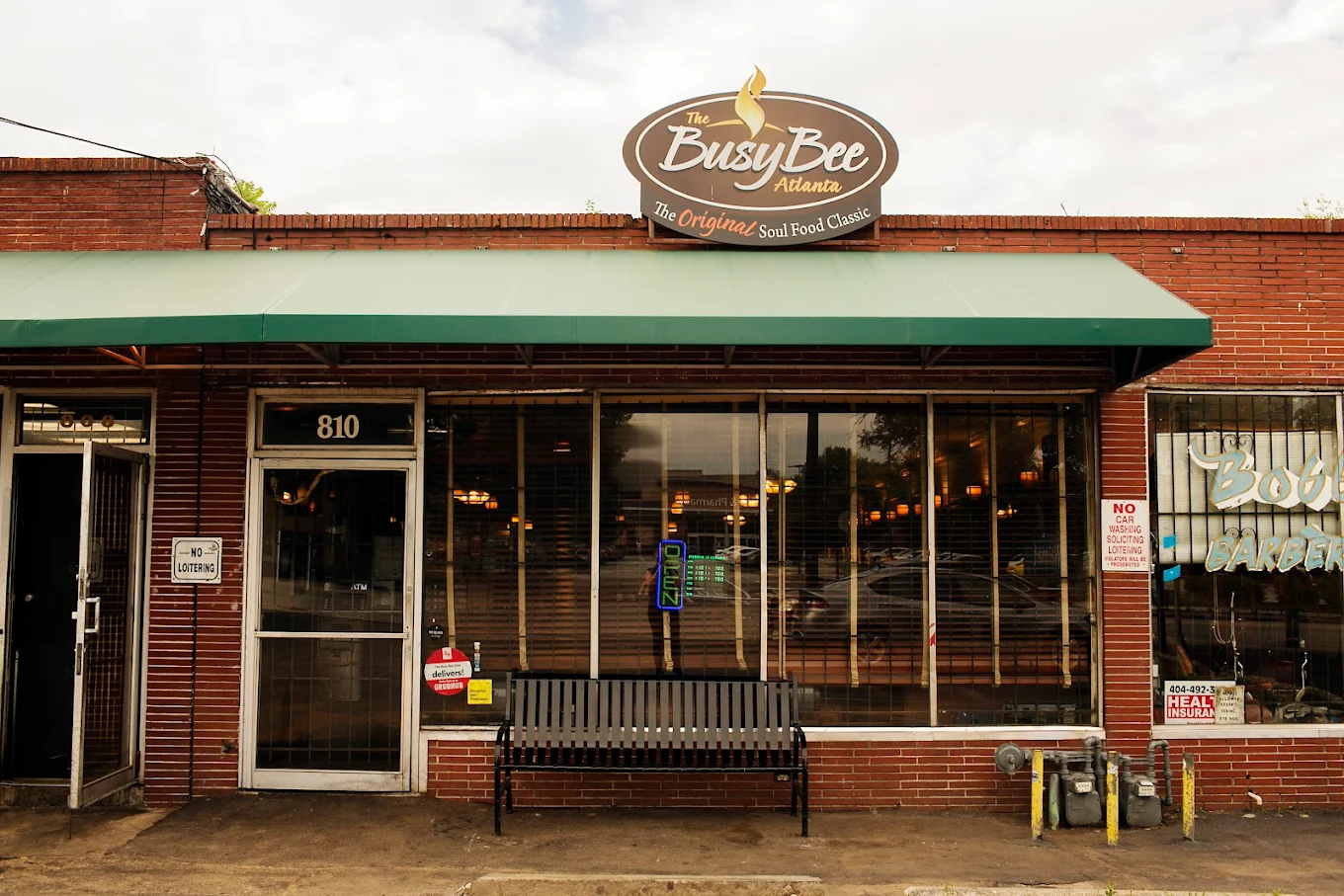 Exterior of The Busy Bee Atlanta restaurant, showing a green awning, large front windows, a black bench, and a neon “Open” sign; brick building with the address number 810 above the door.