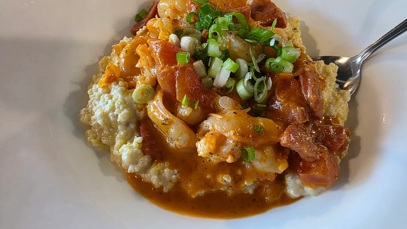 A plate of creamy grits topped with shrimp in a rich, orange-colored sauce, diced tomatoes, and garnished with chopped green onions, served with a spoon.
