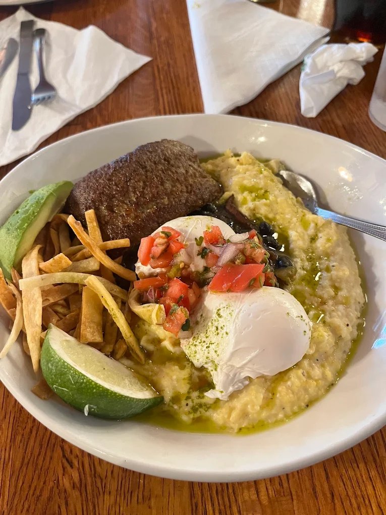 A bowl of creamy grits topped with a poached egg, salsa, grilled beef patty, avocado slices, fried tortilla strips, and a lime wedge, served on a wooden table with utensils and a napkin nearby.