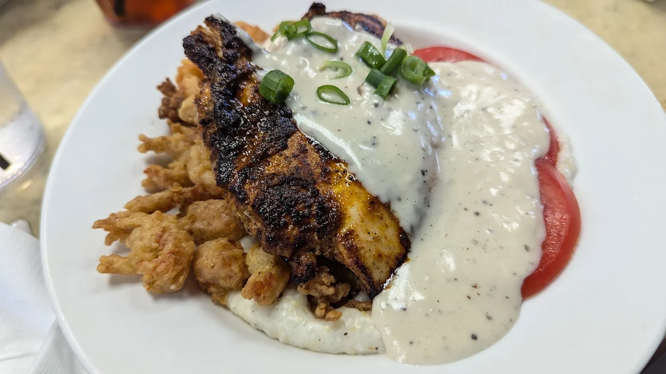 A plate of food featuring blackened fish, fried okra, sliced tomatoes, creamy white gravy with pepper, and chopped green onions, served on a white dish.