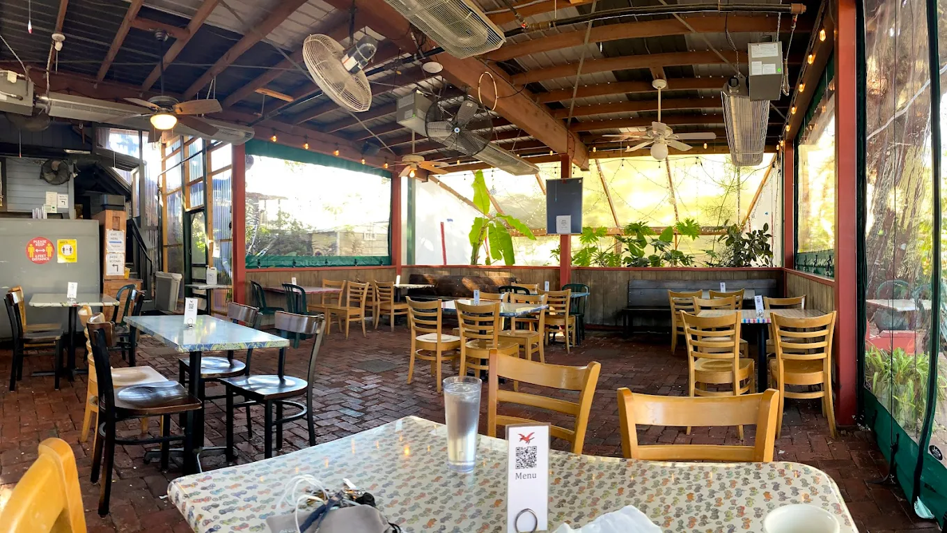 A covered outdoor patio dining area with wooden chairs and tables, brick flooring, ceiling fans, potted plants, and a glass of iced water on the foreground table with a menu and utensils. The restaurant appears quiet and empty.