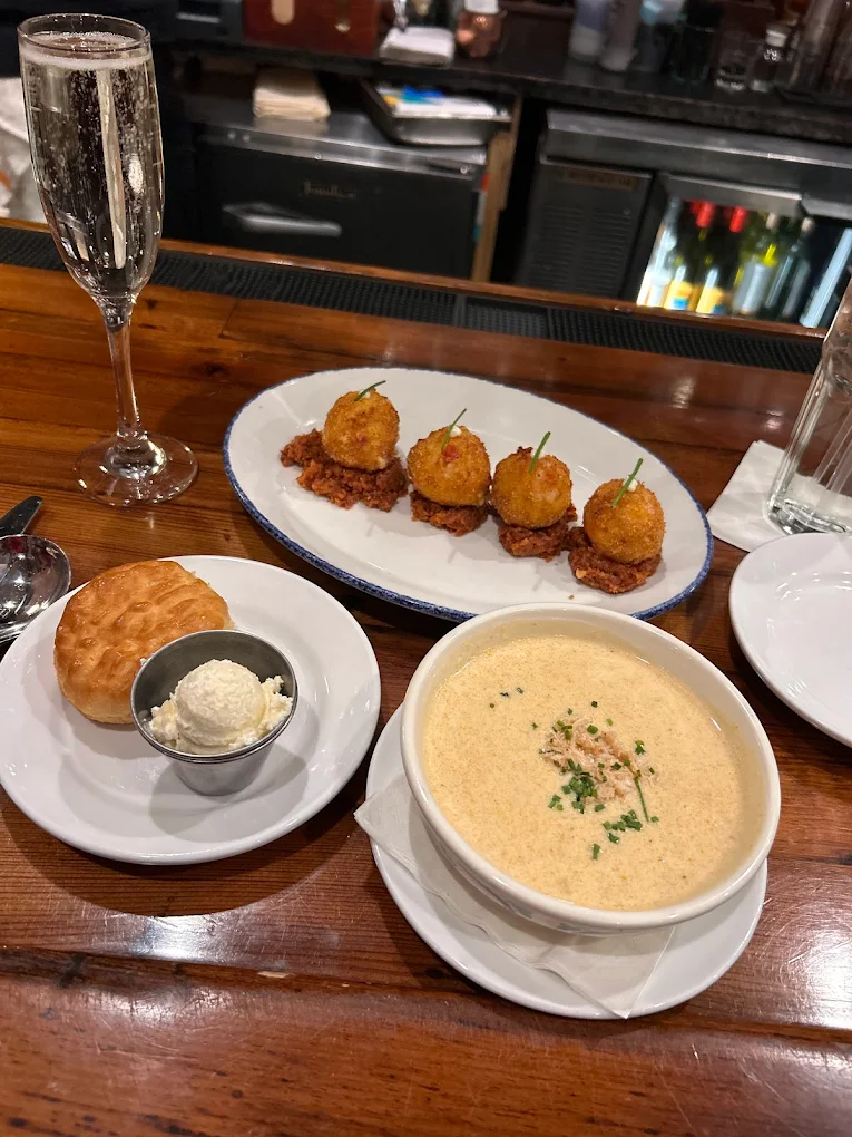 A wooden bar table with a glass of sparkling wine, four fried appetizers on a platter, a bowl of creamy soup topped with chives, and a biscuit with a side of butter on a small plate.