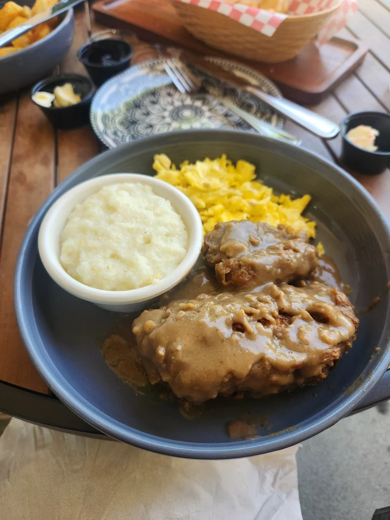 A plate with two pieces of meatloaf topped with brown gravy, a serving of scrambled eggs, and a bowl of creamy grits, set on a wooden table with small containers of butter and bread in the background.