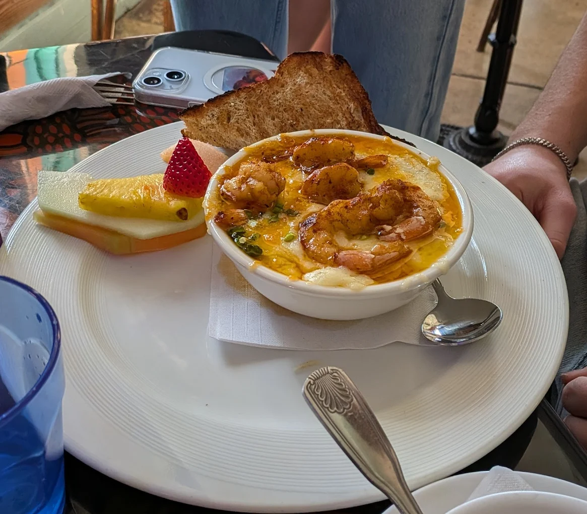 A bowl of shrimp and grits topped with herbs sits on a white plate with toast, a slice of pineapple, cantaloupe, and a strawberry. A spoon, blue glass, and phone are nearby; a person's hands rest by the plate.