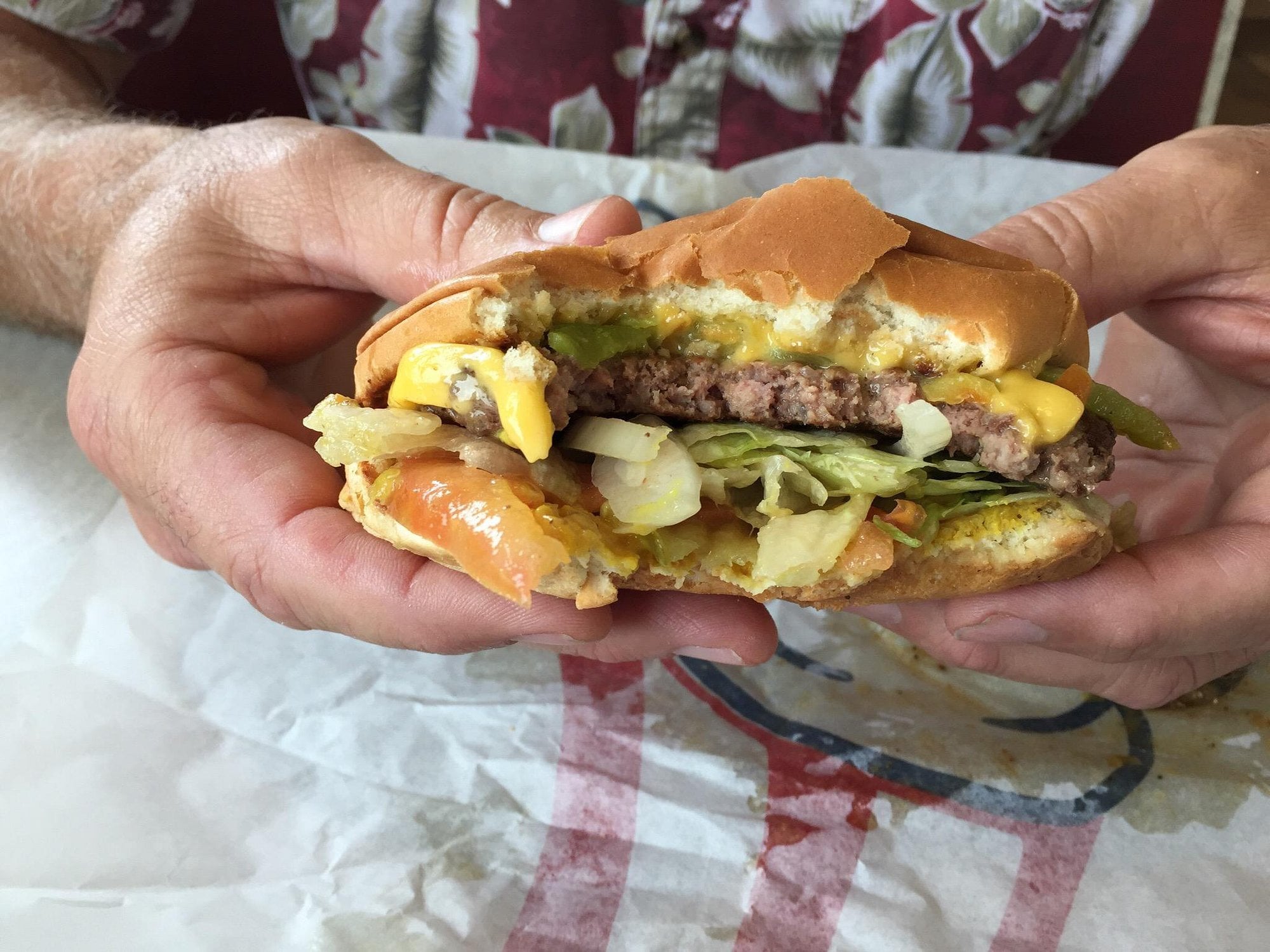 A person holds a partially eaten cheeseburger with lettuce, onions, pickles, and cheese in their hands over a paper wrapper—one of the classic favorites often found at regional fast-food chains.