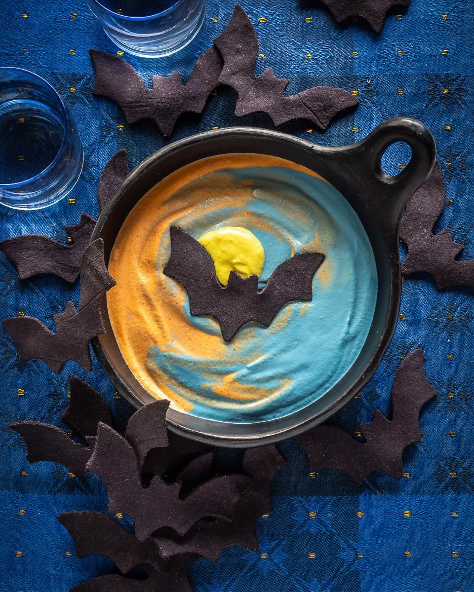 A bowl of colorful dip with orange, yellow, and blue swirls is topped with a bat-shaped cookie. More bat cookies are scattered around on a blue patterned tablecloth with two empty glasses nearby.