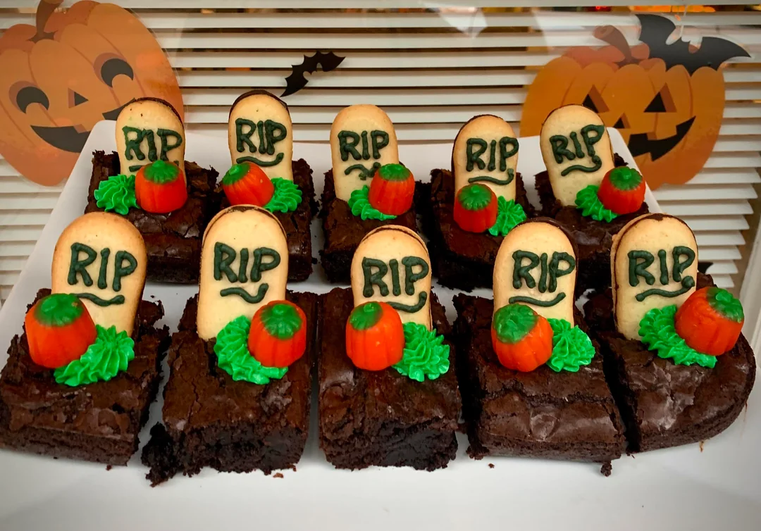 Brownie squares decorated with cookie “tombstones” labeled “RIP,” green icing, and small candy pumpkins, arranged on a tray with Halloween pumpkin decorations in the background.