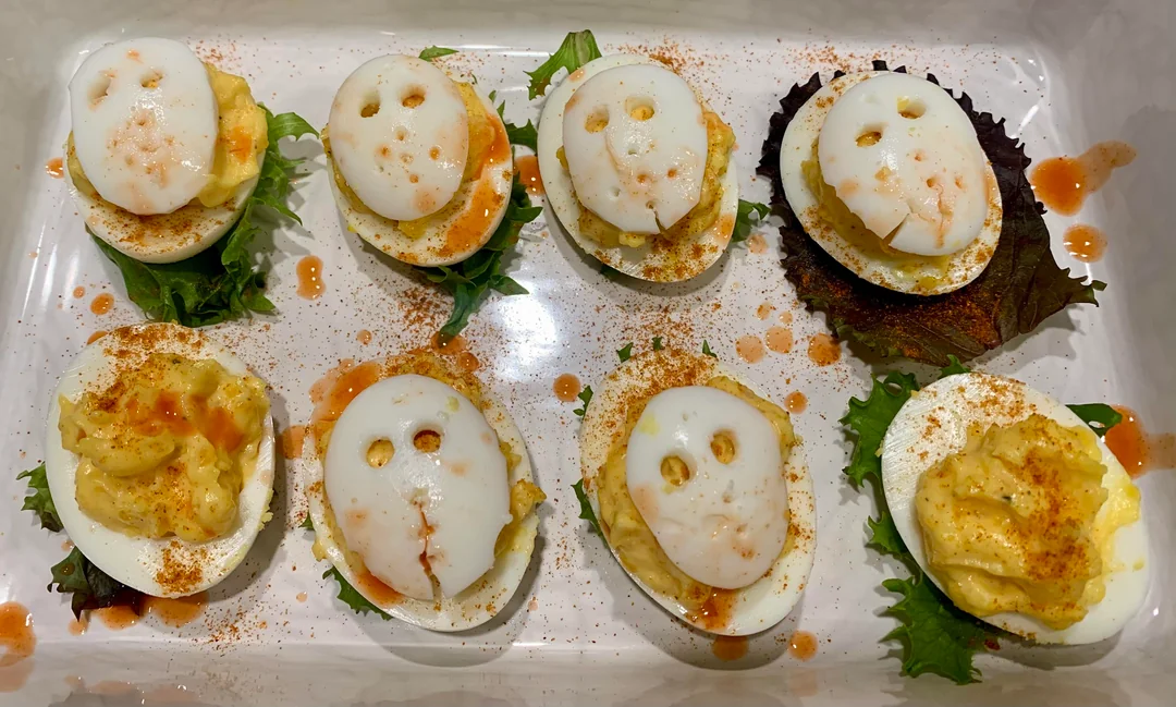Deviled eggs in a tray, some topped with sliced egg whites resembling ghost faces, sprinkled with paprika and hot sauce, on green lettuce leaves.