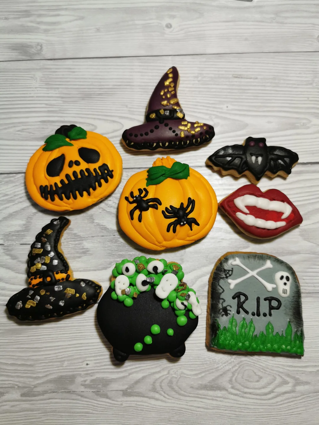 Assorted Halloween-themed cookies shaped like pumpkins, witch hats, a bat, vampire lips, a cauldron, and a tombstone with "RIP," all decorated with colorful icing on a wooden surface.