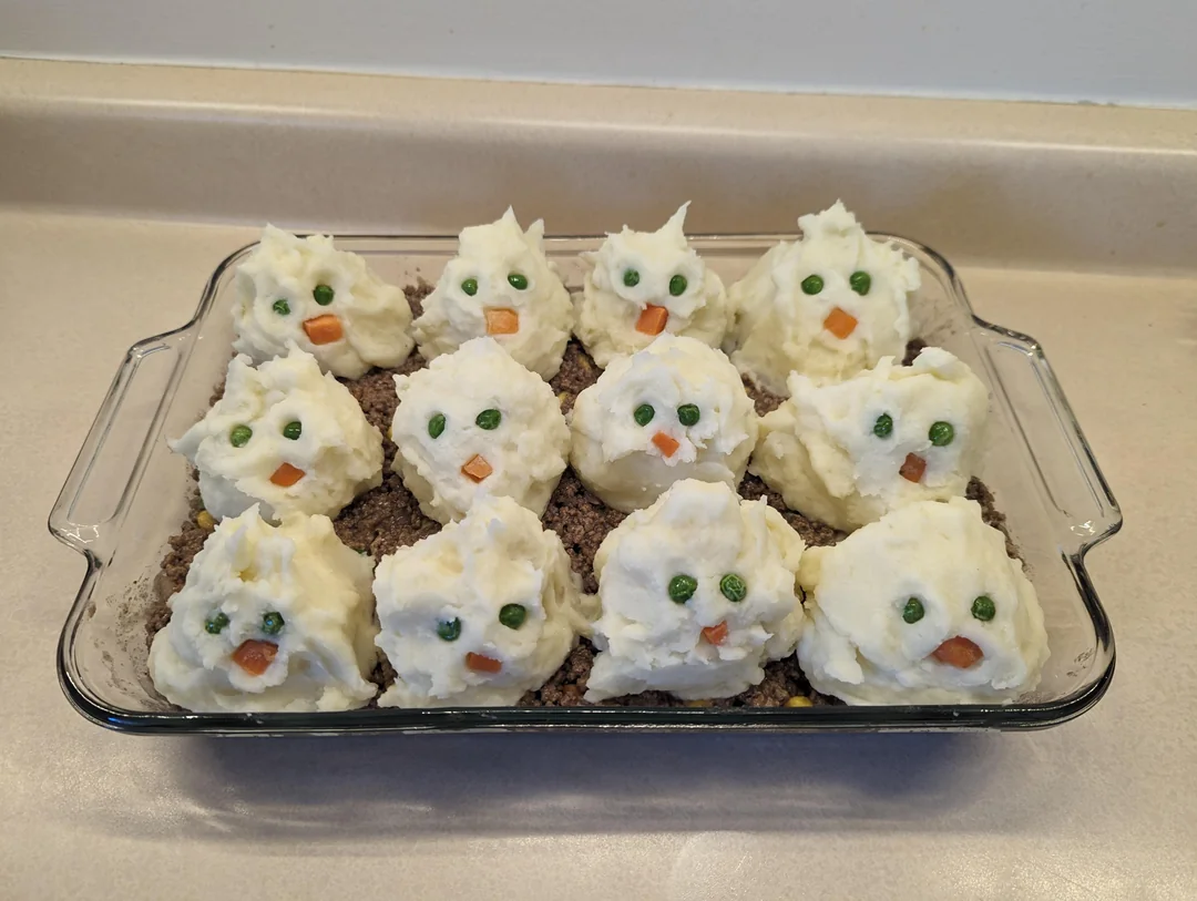 A casserole topped with mashed potato scoops decorated as chicks using peas for eyes and carrot pieces for beaks, arranged in a glass dish.