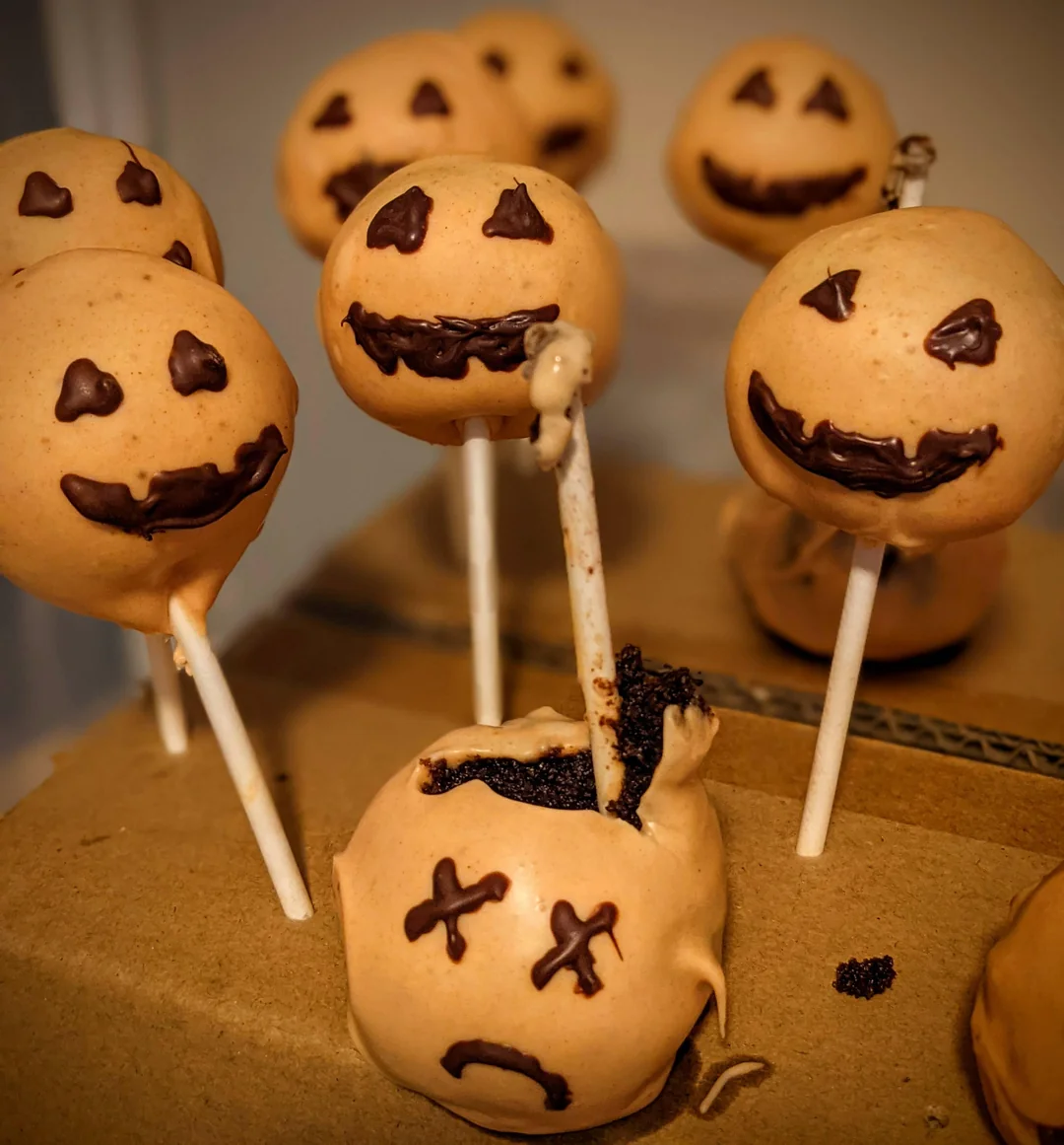 Cake pops decorated as jack-o'-lanterns with orange coating and chocolate faces. One cake pop is broken and decorated with X eyes and a sad mouth, appearing "dead." All are displayed on a cardboard surface.