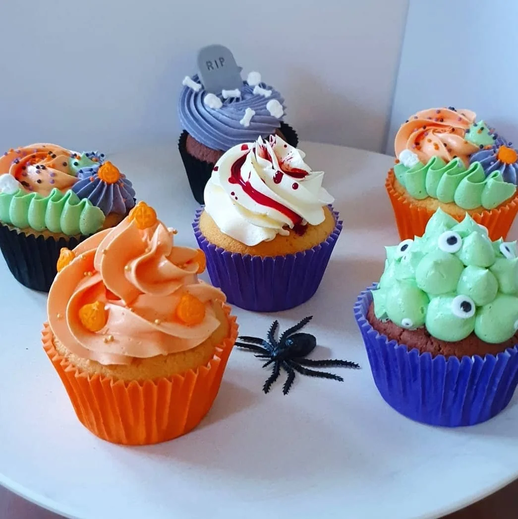 Six colorful Halloween-themed cupcakes with various frosting designs and decorations, including candy eyeballs, a gravestone topper, and a plastic spider on the white plate.