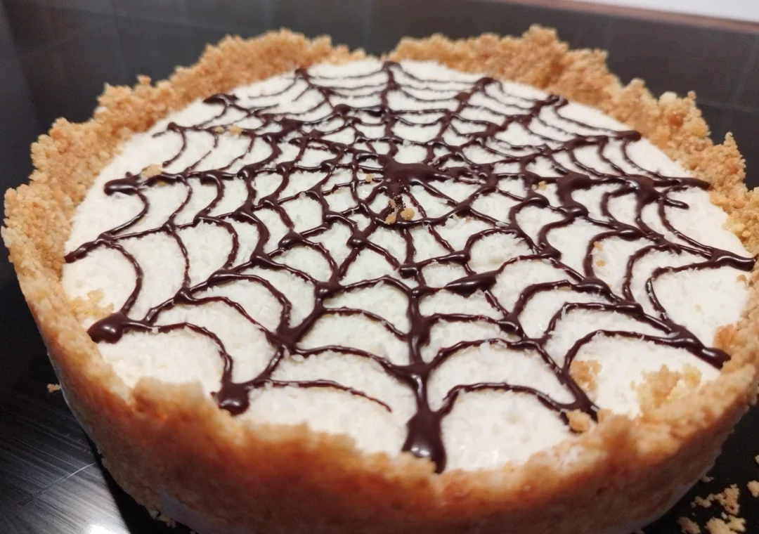 A cheesecake with a crumbly crust and a chocolate web pattern on top, displayed on a dark surface.