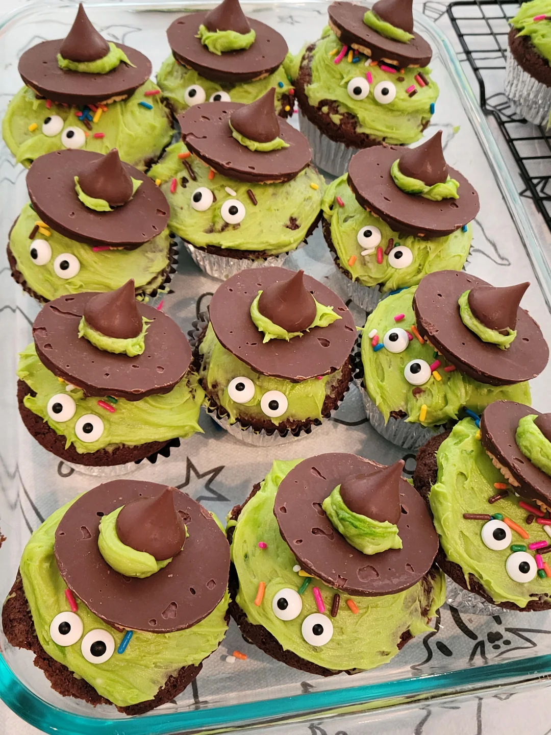 Chocolate cupcakes with green frosting decorated as witches, with candy eyes, chocolate hats, and colorful sprinkles, arranged on a tray and cooling rack.