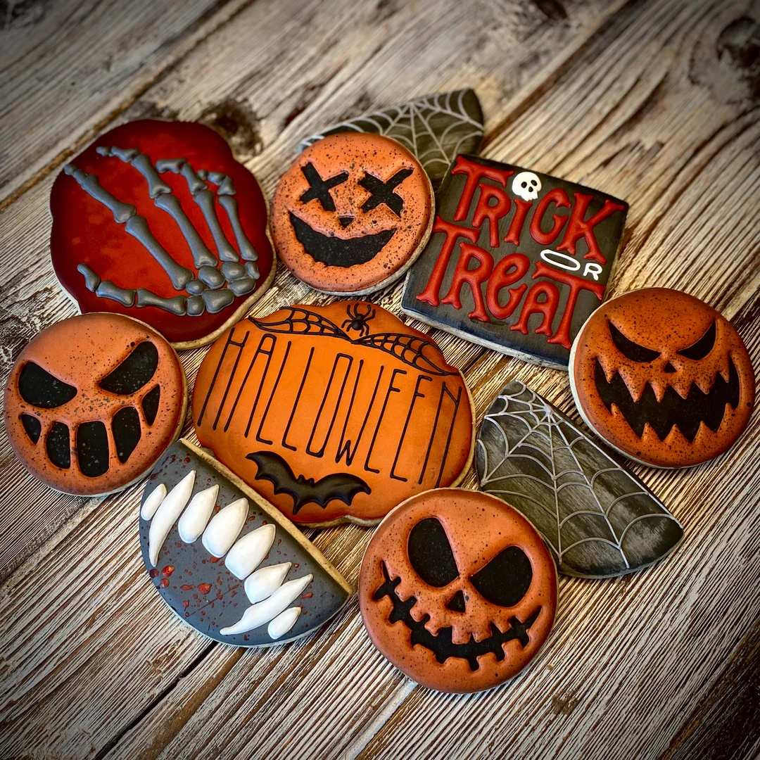 Assorted Halloween-themed cookies shaped and decorated like pumpkins, fangs, skeleton hands, spider webs, and signs with "Trick or Treat" and "Halloween" on a wooden surface.