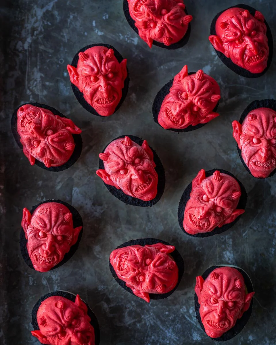 Chocolate cupcakes topped with detailed, scary red devil faces made of icing, arranged on a dark tray.