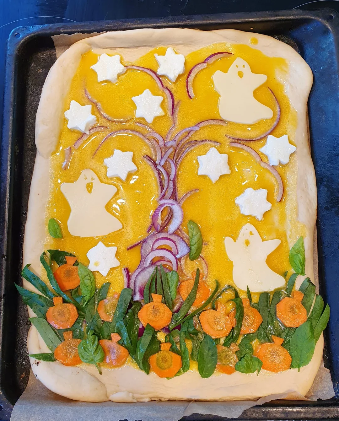 Rectangular pizza with a yellow cheese base, topped with ghost and star-shaped cheese, red onion swirls, spinach, and carrot slices arranged like flowers at the bottom, resembling a whimsical Halloween scene.