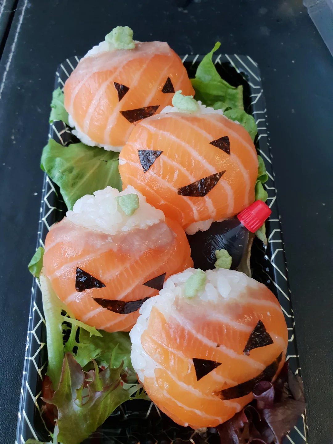Four pieces of sushi shaped like jack-o’-lanterns with salmon slices, nori faces, and wasabi on top, arranged on leafy greens in a black tray with a small soy sauce bottle.