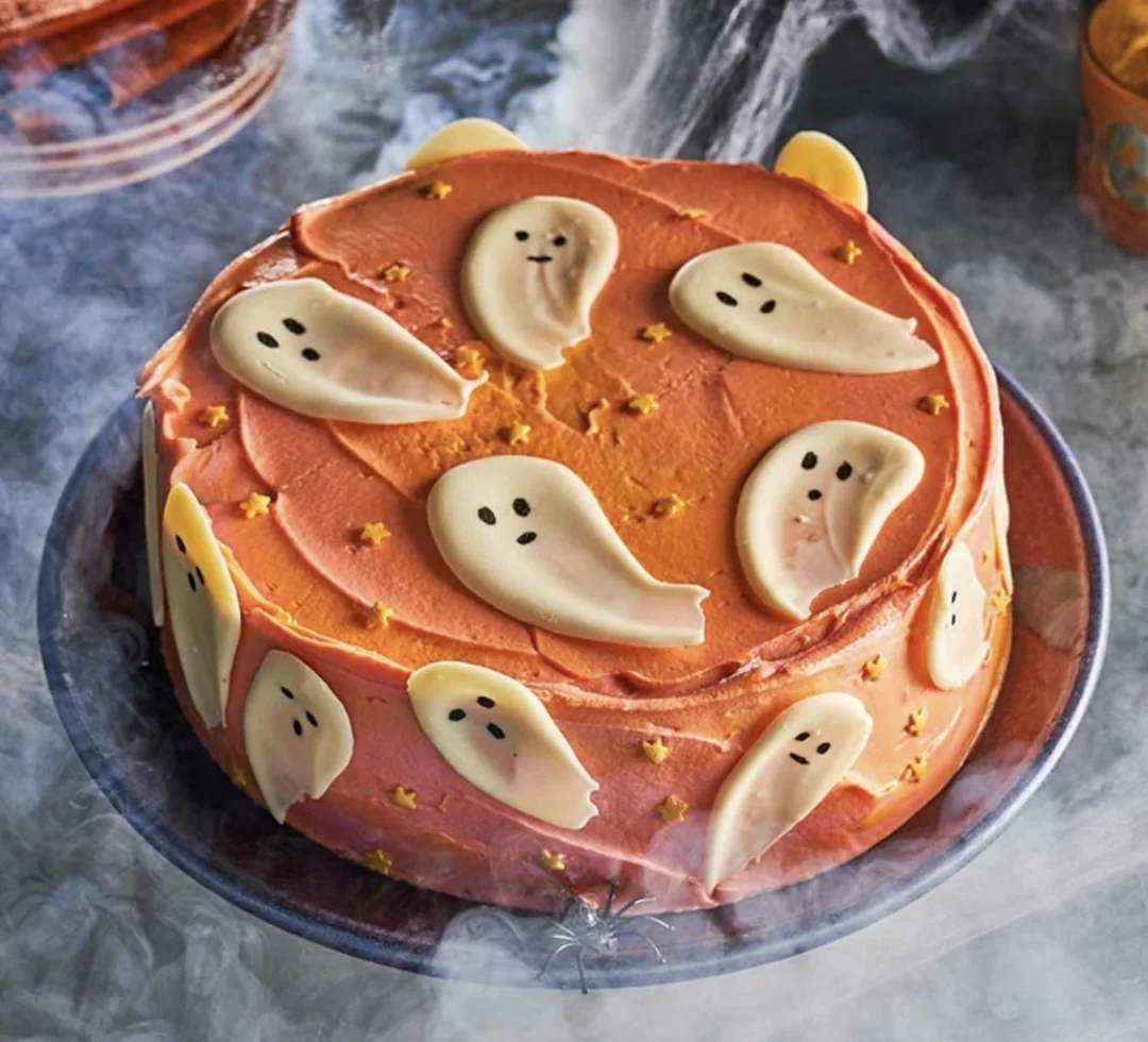 A round cake with orange frosting, decorated with white chocolate ghost shapes and small yellow stars, sits on a plate surrounded by fog for a Halloween-themed presentation.