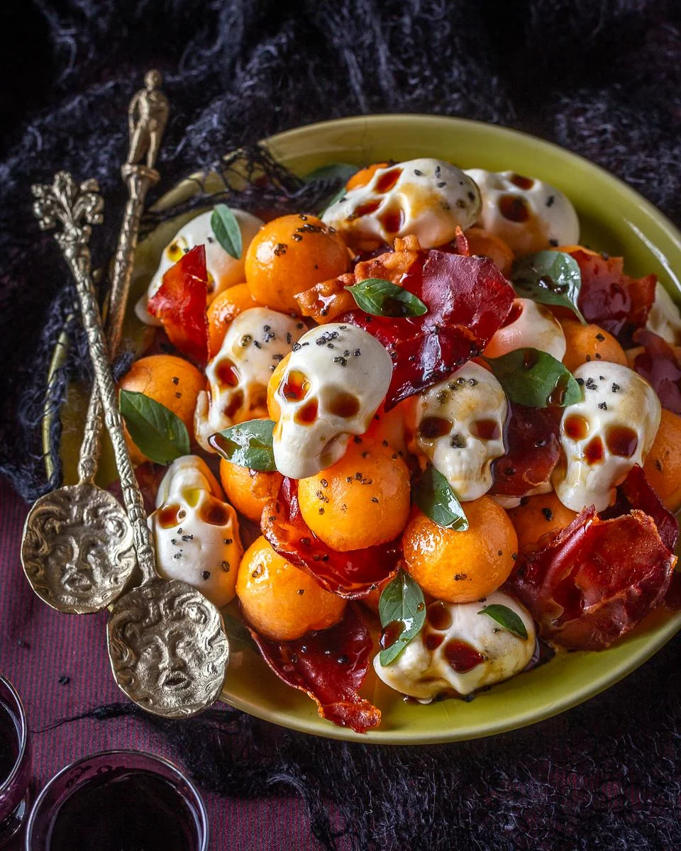 A plate of melon balls, crispy prosciutto, and cheese molded into skull shapes, garnished with herbs. Two ornate gold spoons with face designs rest beside the dish. Homemade Halloween treats. 