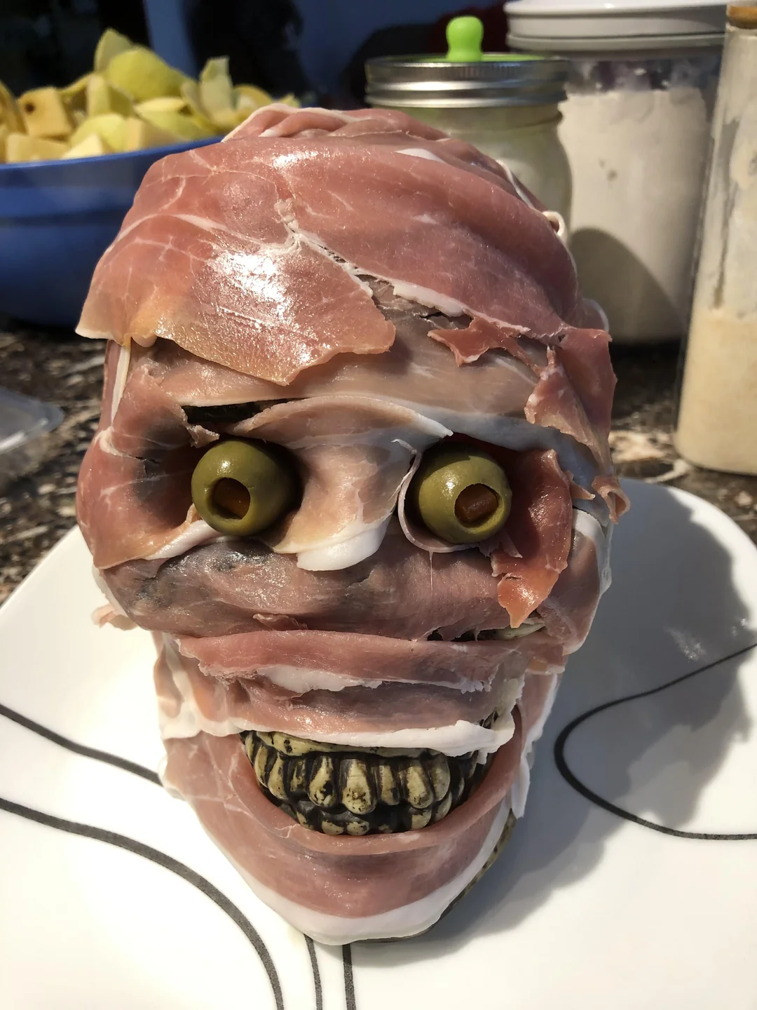 A skull-shaped food platter wrapped in deli meat with olives for eyes and green peppers for teeth, arranged to resemble a creepy mummy face on a white plate.