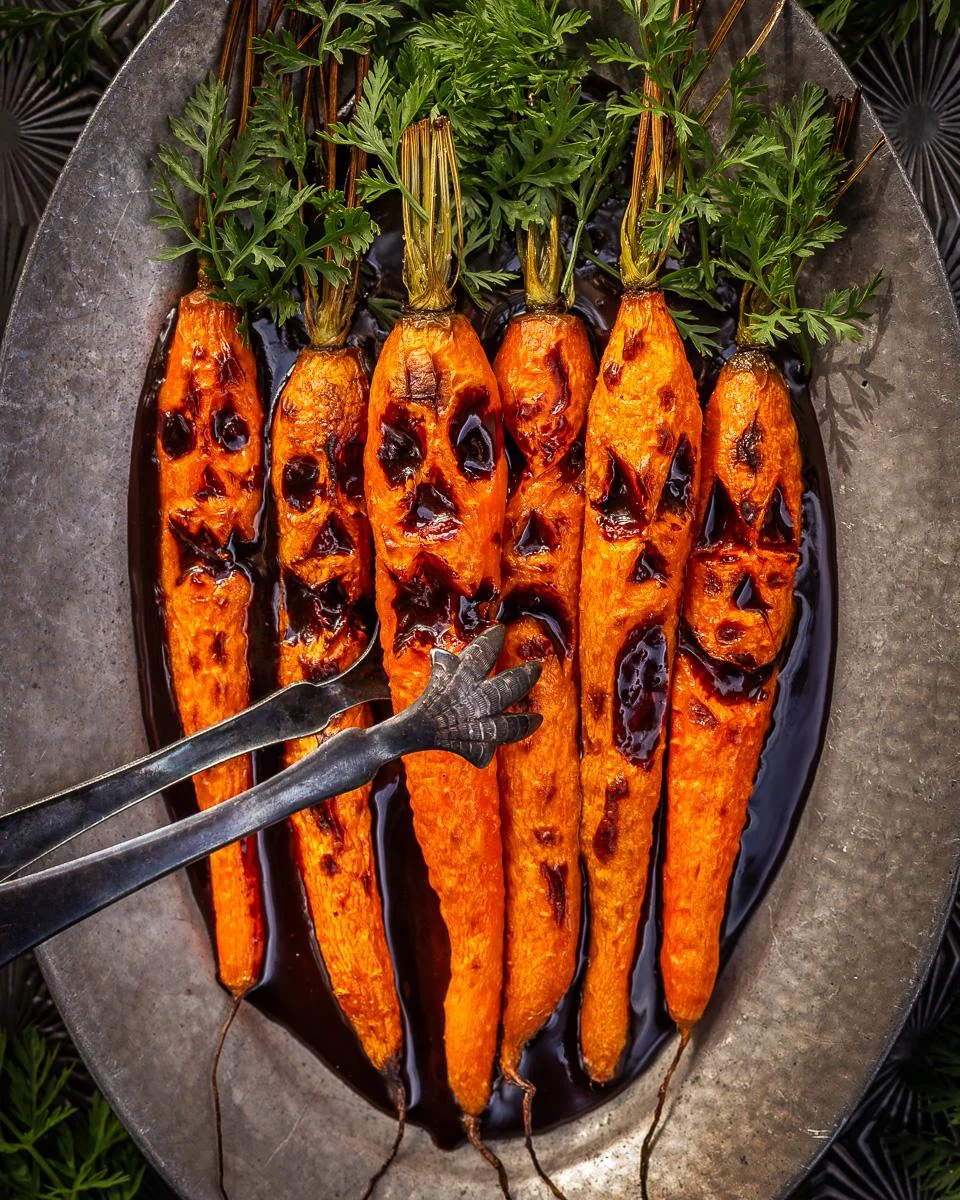 Roasted carrots with carved spooky faces are arranged on a platter, resembling Halloween jack-o’-lanterns. A pair of tongs shaped like a skeletal hand is grabbing one carrot.