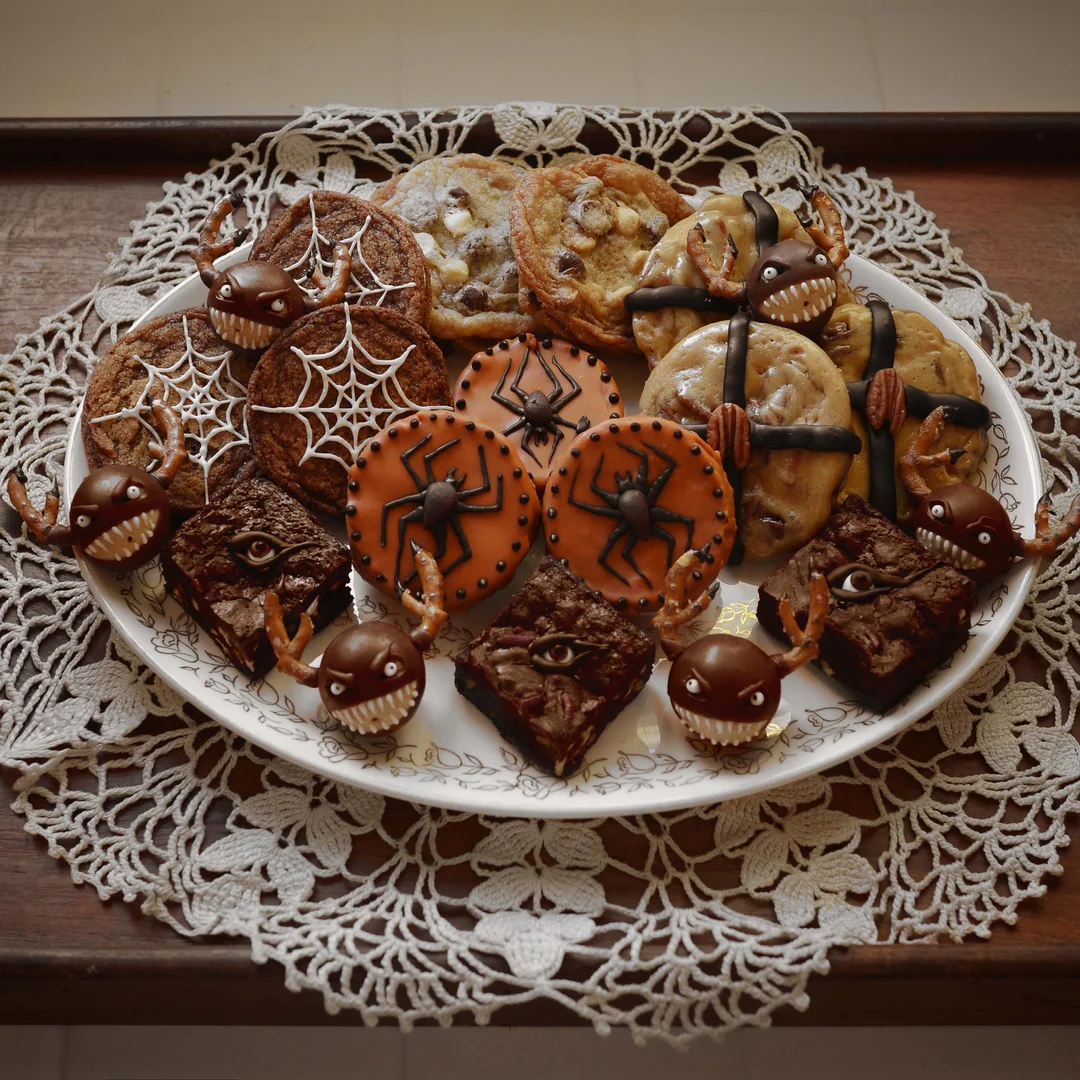 A plate of Halloween-themed cookies and brownies, decorated with spider webs, spiders, eyes, and pecans, sits on a lace doily on a wooden surface.