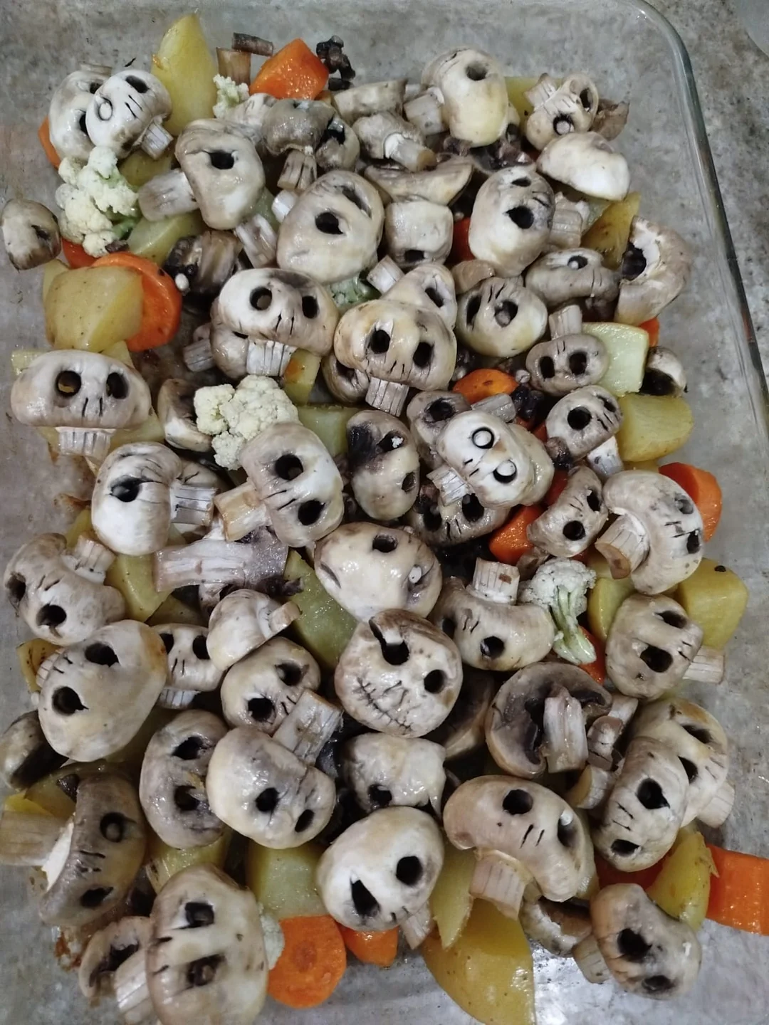 Raw mushrooms with faces resembling skulls are arranged on top of sliced potatoes, carrots, and cauliflower in a glass baking dish, creating a Halloween-themed vegetable display.