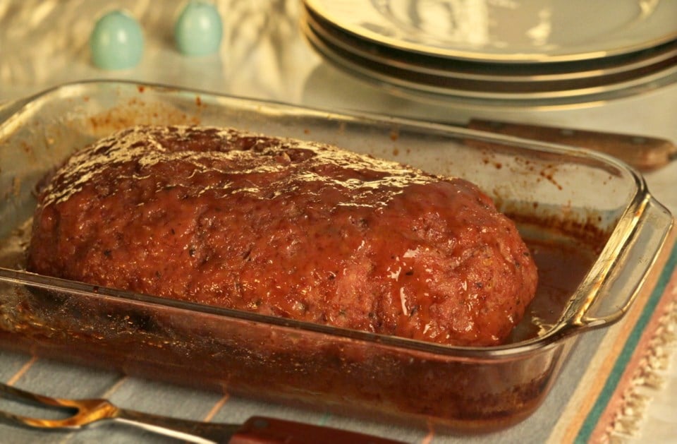 A glazed meatloaf sits in a glass baking dish on a table, with plates and utensils in the background.