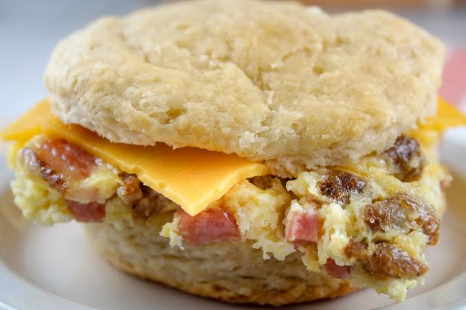 A close-up of a breakfast biscuit sandwich filled with scrambled eggs, sausage, ham, and a slice of cheddar cheese on a white plate.