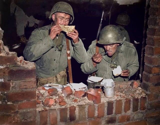 Two soldiers in uniform and helmets eat food while standing behind a damaged brick wall. One drinks from a metal cup, the other eats bread. Metal dishes and debris are visible on the wall in front of them, evoking a sense of nostalgic dinners shared amid the hardships of war.