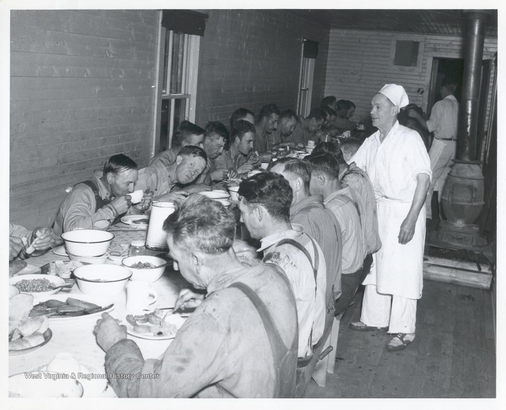 A group of men sit closely at a long table eating a meal, while a man in a white uniform stands beside them. The setting appears to be a cafeteria or mess hall with wooden walls and a wood stove, evoking the warmth and camaraderie of nostalgic dinners shared in simpler times.